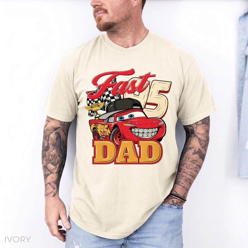 May include: An ivory short-sleeve t-shirt featuring a graphic of a red race car wearing a black baseball cap and braces. The design includes a checkered flag, the number 95, and the red and yellow text "Fast DAD" printed on the front.