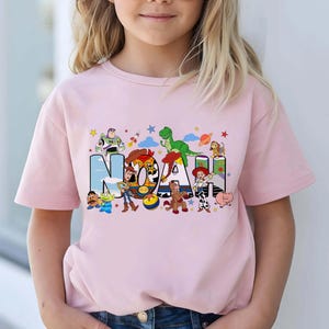 K&ouml;nnte beinhalten: Ein hellrosa T-Shirt mit kurzen &Auml;rmeln und einem grafischen Aufdruck des Namens NOAH in gro&szlig;en Blockbuchstaben. Die Buchstaben sind mit verschiedenen bunten Zeichentrickfiguren gef&uuml;llt, darunter ein Weltraum-Ranger, ein Cowboy und ein Dinosaurier.