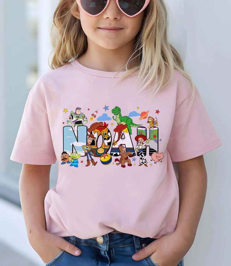 K&ouml;nnte beinhalten: Ein hellrosa T-Shirt mit kurzen &Auml;rmeln und einem grafischen Aufdruck des Namens NOAH in gro&szlig;en Blockbuchstaben. Die Buchstaben sind mit verschiedenen bunten Zeichentrickfiguren gef&uuml;llt, darunter ein Weltraum-Ranger, ein Cowboy und ein Dinosaurier.