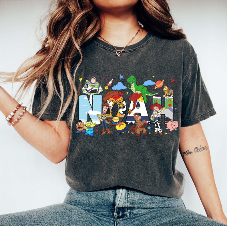 K&ouml;nnte beinhalten: Ein anthrazitfarbenes Kurzarm-T-Shirt mit dem Namen NOAH in gro&szlig;en Blockbuchstaben. Das Design zeigt bunte Zeichentrickfiguren aus der Toy Story-Reihe, darunter Woody, Buzz Lightyear, Jessie, Rex und Hamm, sowie kleine Sterne.