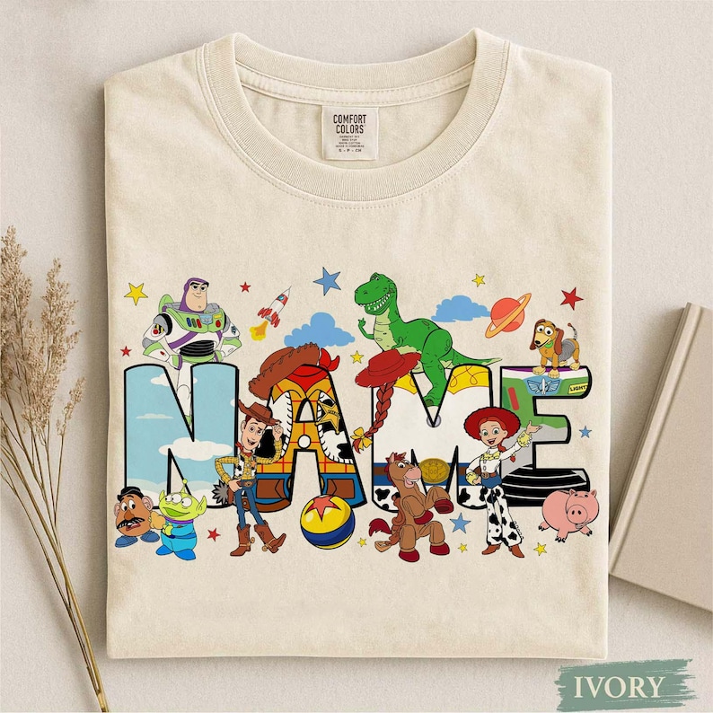 K&ouml;nnte beinhalten: Ein elfenbeinfarbenes T-Shirt mit Rundhalsausschnitt und einem Grafikdruck verschiedener Toy Story-Figuren, die das Wort NAME in gro&szlig;en Blockbuchstaben umgeben. Das Design zeigt Buzz Lightyear, Woody, Rex, Jessie, Slinky Dog und weitere.