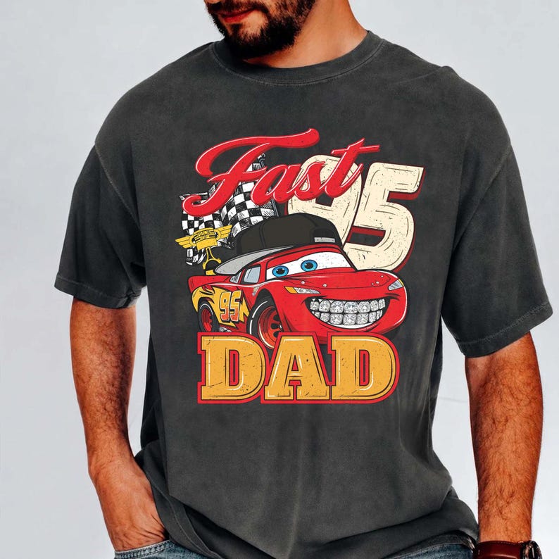 May include: A charcoal gray short-sleeve t-shirt featuring a graphic of a red race car wearing a black baseball cap and braces. The design includes a checkered flag, the number 95, and the text "Fast 95 DAD" in bold red and yellow lettering.