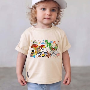 K&ouml;nnte beinhalten: Ein cremefarbenes Kurzarm-T-Shirt mit einem bunten Grafikdruck des Namens JAX. Die Buchstaben sind mit verschiedenen animierten Spielzeugfiguren verziert, darunter ein Cowboy, ein Weltraum-Ranger, ein gr&uuml;ner Dinosaurier und ein Cowgirl.