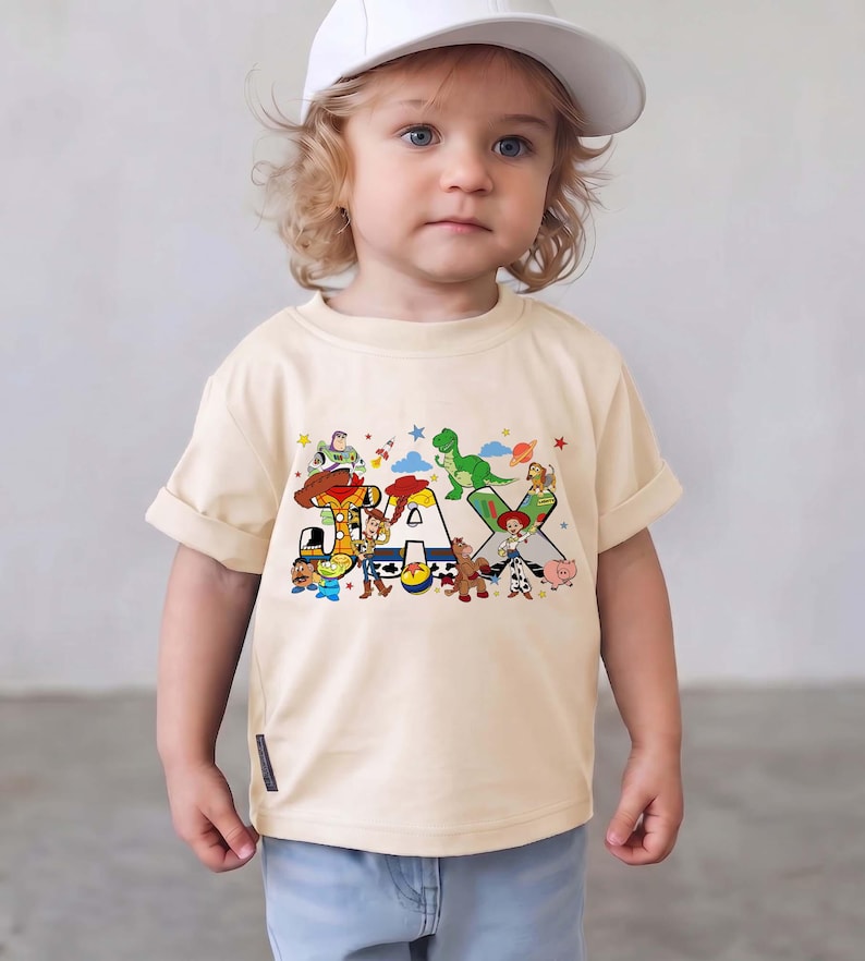 K&ouml;nnte beinhalten: Ein cremefarbenes Kurzarm-T-Shirt mit einem bunten Grafikdruck des Namens JAX. Die Buchstaben sind mit verschiedenen animierten Spielzeugfiguren verziert, darunter ein Cowboy, ein Weltraum-Ranger, ein gr&uuml;ner Dinosaurier und ein Cowgirl.