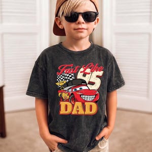 May include: A charcoal grey acid-wash t-shirt featuring a graphic of a red race car with a black cap and braces. The text reads Fast Like 95 DAD, accompanied by a checkered flag. The shirt has a crew neckline and short sleeves, showcasing a vintage-inspired wash.