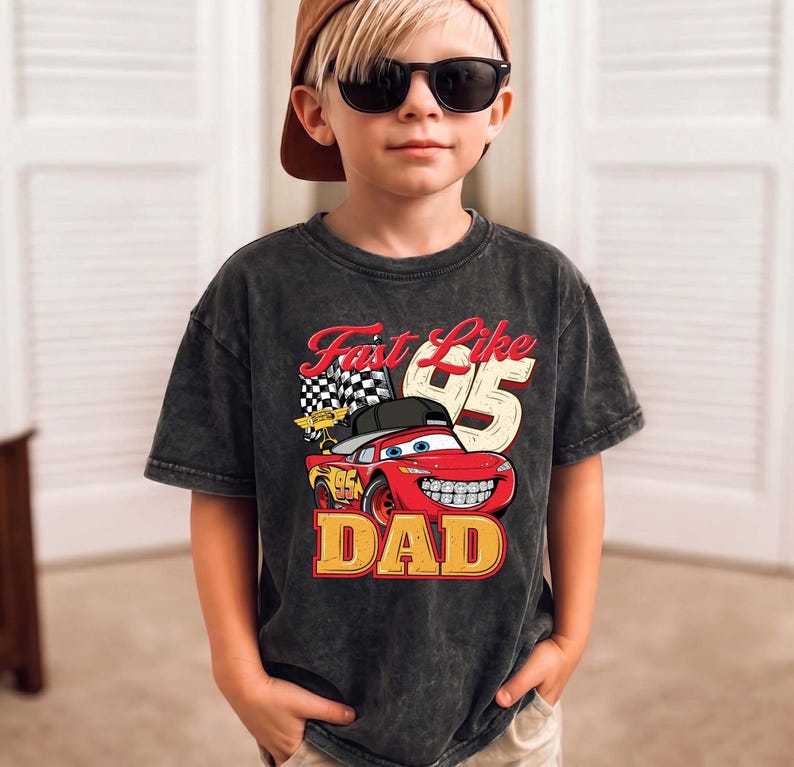 May include: A charcoal grey acid-wash t-shirt featuring a graphic of a red race car with a black cap and braces. The text reads Fast Like 95 DAD, accompanied by a checkered flag. The shirt has a crew neckline and short sleeves, showcasing a vintage-inspired wash.