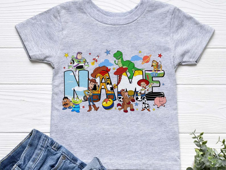 K&ouml;nnte beinhalten: Ein kurz&auml;rmeliges T-Shirt in Grau meliert mit einem gro&szlig;en Grafikdruck des Wortes NAME in Blockbuchstaben. Die Buchstaben sind mit verschiedenen Zeichentrickfiguren verziert, darunter ein Cowboy, ein Weltraum-Ranger, ein Dinosaurier und ein Cowgirl.