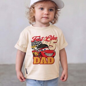 May include: A cream-colored short-sleeve t-shirt featuring a graphic of a red race car wearing a black baseball cap. The text reads Fast Like 95 DAD, accompanied by a checkered flag and the number 95 in a large, bold, tan-colored font.