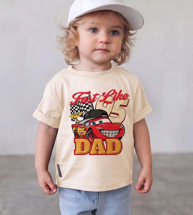 May include: A cream-colored short-sleeve t-shirt featuring a graphic of a red race car wearing a black baseball cap. The text reads Fast Like 95 DAD, accompanied by a checkered flag and the number 95 in a large, bold, tan-colored font.