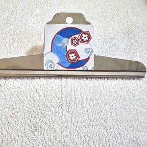 May include: A silver metal document clip featuring a hand-painted design on the front. The artwork displays a blue crescent moon shape accented with red and white floral motifs and light blue cloud-like swirls against a white background.