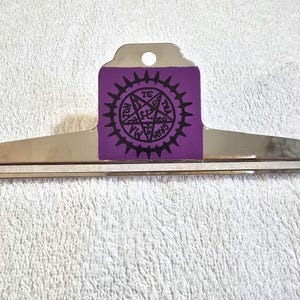 May include: A silver metal bulldog clip featuring a purple rectangular insert on the handle. The insert displays a black circular emblem with a pentagram design and the text "MATON GRAM" printed in a circular pattern around the center.