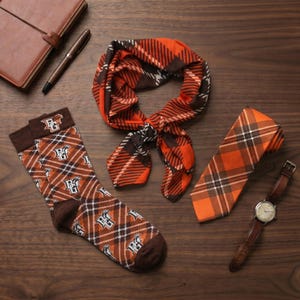 May include: An assortment of accessories in a plaid pattern. Includes a neck scarf, tie, and socks in orange, brown, and white. A brown leather watch and a pen and notebook are also present on a wooden surface.
