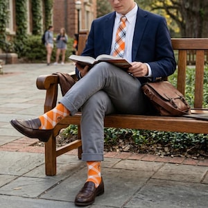 May include: A person seated on a wooden bench, wearing a navy blazer, gray pants, and orange patterned socks. The socks feature a diamond pattern with a paw print. Brown leather shoes and a matching satchel complete the look. The person is reading a book.