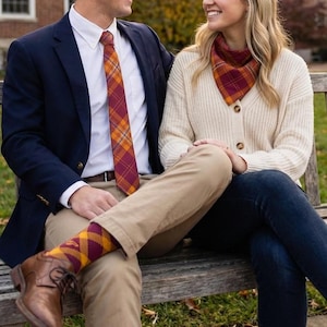 May include: A person wearing a navy blazer, white shirt, and plaid tie in burgundy and gold. The person is wearing khaki pants and burgundy and gold plaid socks. A person wearing a cream cardigan and a matching plaid scarf is also in the image.
