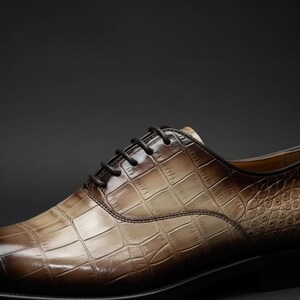 May include: A close-up of a brown leather dress shoe with a crocodile skin pattern. The shoe has black laces and a dark sole. The leather has a gradient effect, transitioning from light to dark brown.