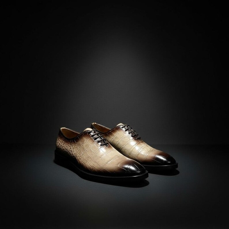 May include: A pair of brown and tan leather dress shoes with a textured, crocodile-like pattern. The shoes have black laces and a dark brown toe. The background is black.