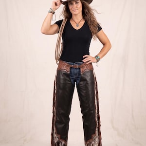 Peut inclure: Une personne portant un chapeau de cowboy marron, un t-shirt noir à col en V et un jean. Elle porte des chaps en cuir marron avec des franges et une ceinture décorative. Une corde est drapée sur son épaule. La marque "LEATHERLEAVESCO." est en haut à gauche.