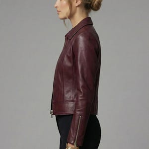 May include: A burgundy leather jacket with a zipper closure and a collared neckline. The jacket has a tailored fit and long sleeves with zippered cuffs. The model is wearing black pants and gold bracelets. The logo "LEATHER LEAVES PVT LTD" is visible.