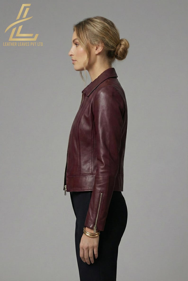 May include: A burgundy leather jacket with a zipper closure and a collared neckline. The jacket has a tailored fit and long sleeves with zippered cuffs. The model is wearing black pants and gold bracelets. The logo "LEATHER LEAVES PVT LTD" is visible.