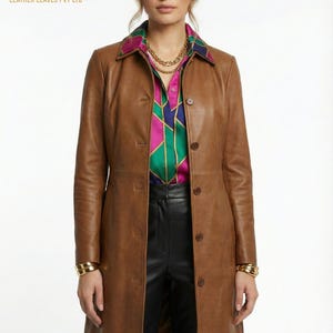 Könnte beinhalten: Ein brauner Ledercoat mit Knopfleiste. Der Mantel wird über einem bunten Hemd und einer schwarzen Hose getragen. Goldarmbänder und eine goldene Halskette sind sichtbar. Das Logo "LEATHER LEAVES PVT LTD" ist zu sehen.