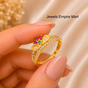 Könnte beinhalten: Ein goldener Ring mit einem kronenförmigen Oberteil, das mit rosafarbenen Steinen im Marquise-Schliff und einem zentralen grünen Stein verziert ist, sowie einem Band, das mit kleinen klaren Steinen besetzt ist. Der Text Jewels Empire Mart ist sichtbar.