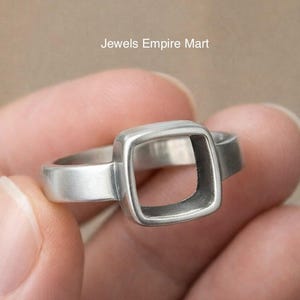 Könnte beinhalten: Ein silberner Ring mit einem quadratischen Design mit offenem Zentrum. Der Ring wird in der Hand einer Person gehalten, wodurch seine geometrische Form und das metallische Finish hervorgehoben werden. Der Text "Jewels Empire Mart" ist im Hintergrund sichtbar.