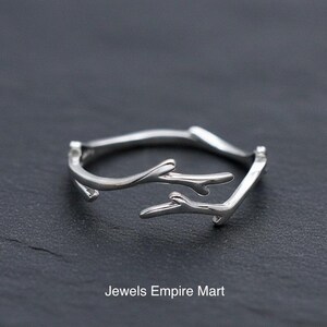 May include: A silver-colored ring with a twig-like design. The ring features a branch-like structure with multiple prongs, creating a nature-inspired aesthetic. The ring is displayed on a dark surface. Text on the image reads "Jewels Empire Mart."