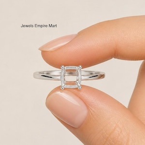 Könnte beinhalten: Ein silberner Ring mit quadratischer Fassung, gehalten zwischen Fingern. Der Ring hat einen dünnen Ring und vier kleine Zinken an jeder Ecke der Fassung. Der Text "Jewels Empire Mart" ist in der oberen linken Ecke sichtbar.