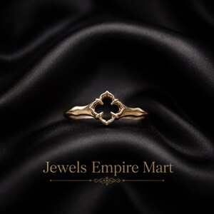 Könnte beinhalten: Ein goldener Ring mit einem mittigen, vierblättrigen Ausschnitt mit spitzen Kanten. Die Ringschiene hat eine glatte, zulaufende Oberfläche. Unter dem Ring steht der Text Jewels Empire Mart in einer Serifenschrift mit einem Zierelement.