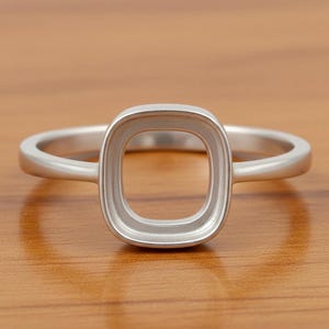 Könnte beinhalten: Ein silberner Ring mit quadratischer Lünettenfassung. Der Ring hat ein schlichtes Band und eine quadratische Fassung, die dazu bestimmt ist, einen Edelstein oder ein anderes dekoratives Element zu halten. Der Ring wird auf einer Holzoberfläche gezeigt.