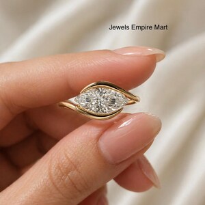 May include: A gold ring featuring a marquise-cut diamond. The diamond is set in a unique, flowing design. The ring is held against a soft, white background. The text "Jewels Empire Mart" is visible.