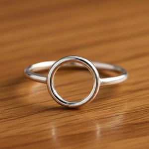 Op de afbeelding: Een zilveren ring met een cirkelvormig ontwerp. De ring heeft een gepolijste, open cirkel als hoofdelement, gezet op een dunne band. De ring wordt gepresenteerd op een houten oppervlak, wat de minimalistische stijl benadrukt.