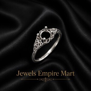 Könnte beinhalten: Eine silberne Ringfassung mit einem komplizierten durchbrochenen Design, einer zentralen runden Halterung für einen Edelstein und vier Krappen. Das Band zeigt detaillierte Knotenmuster auf beiden Seiten und bildet einen dekorativen Rahmen.