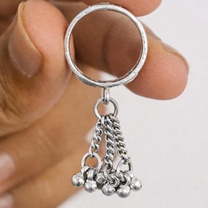 Könnte beinhalten: Ein silberner Ring mit einer daran hängenden Kette aus kleinen, runden, silbernen Perlen. Der Ring wird zwischen den Fingern vor einem schlichten Hintergrund gehalten. Das Band des Rings hat ein strukturiertes Design.