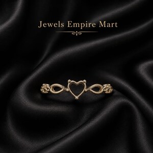Könnte beinhalten: Ein goldfarbener Ring mit einer zentralen herzförmigen Fassung, flankiert von unendlichkeitsartigen Schleifen und strukturierten Seitenakzenten. Der Text Jewels Empire Mart steht über dem Ring, der auf einem dunklen, drapierten Stoff liegt.