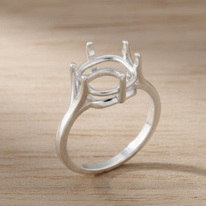 Puede incluir: Un engaste de anillo plateado con un centro ovalado y cuatro garras. La banda del anillo se curva con gracia, y el engaste está diseñado para sostener una piedra preciosa. El anillo se muestra sobre una superficie de madera clara.