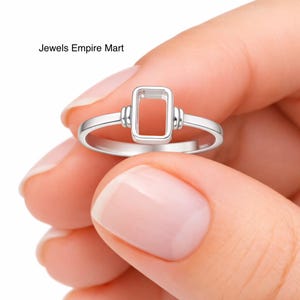 May include: A silver ring with a rectangular setting. The ring has a simple band and a rectangular frame for a gemstone. The ring is held in a hand. The text "Jewels Empire Mart" is in the upper left corner.