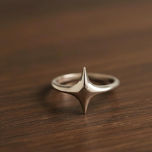 Könnte beinhalten: Ein silberner Ring mit Sternenmuster. Der Ring hat einen zentralen, vierzackigen Stern, der sich aus dem Band erhebt. Der Ring ist auf einer dunklen Holzoberfläche platziert, was seine polierte, reflektierende Oberfläche hervorhebt.