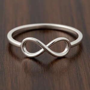 May include: A silver-colored ring featuring an infinity symbol design. The ring is set against a dark brown wooden surface. The infinity symbol is the focal point, with a smooth, polished finish.