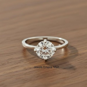 Könnte beinhalten: Ein silberner Ring mit einem großen, runden, klaren Edelstein. Der Ring liegt auf einer Holzoberfläche. Der Text "Jewels Empire Mart" ist unten im Bild zu sehen.