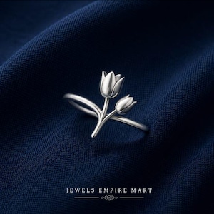Könnte beinhalten: Ein silberfarbener Ring mit einem Design aus zwei Tulpenblüten mit glatten, polierten Blütenblättern und einem schmalen Band. Der Text JEWELS EMPIRE MART ist in Weiß unter dem Ring aufgedruckt, ergänzt durch eine kleine Zierlinie und ein Symbol.