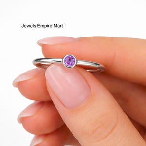 Könnte beinhalten: Ein silberner Ring mit einem runden, lila Edelstein. Der Ring wird in einer Hand mit manikürten Nägeln gehalten. Der Text "Jewels Empire Mart" ist oben links zu sehen.