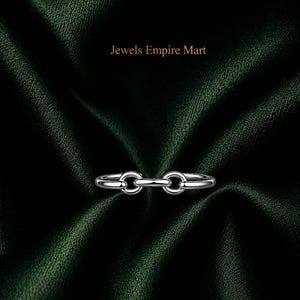 Könnte beinhalten: Ein silberfarbener Ring mit einem Kettenglied-Design, bestehend aus zwei kreisförmigen Schlaufen, die durch einen zentralen horizontalen Stab verbunden sind. Der Text Jewels Empire Mart steht über dem Ring. Das Metall hat eine polierte Oberfläche.
