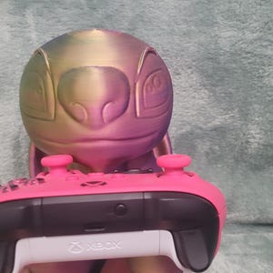 May include: A multi-coloured Stitch-themed controller holder with a pink Xbox controller. The holder is a 3D-printed design of Stitch's head, with the controller resting below. The controller is pink and black.