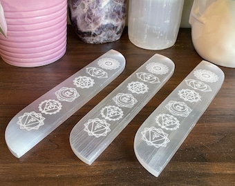 May include: Three white selenite charging plates with etched chakra symbols. The plates are arranged in a row on a wooden surface.