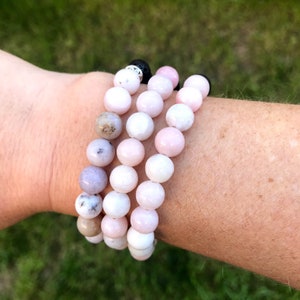May include: Three stacked bracelets made of pink and white gemstone beads. The bracelets are worn on a person's wrist.
