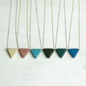 May include: Seven necklaces with triangle-shaped pendants made of lava rock. The pendants are in various colors, including beige, pink, blue, teal, dark green, and dark blue. The necklaces are on a white surface.