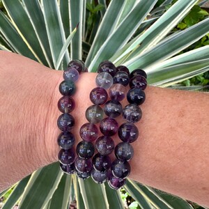 May include: A multi-strand bracelet made of purple, green, and blue fluorite beads. The beads are round and smooth, and the bracelet is stacked on a person's wrist.