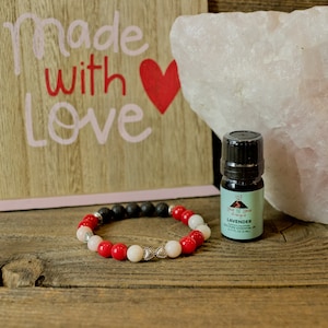 May include: A red, white, and black beaded bracelet with a silver heart charm, a small bottle of lavender essential oil, and a wooden sign that says "Made with Love" with a red heart.