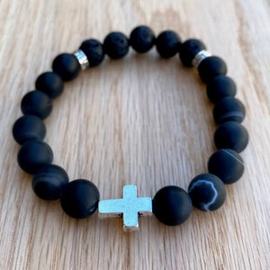 May include: Black beaded bracelet with a silver cross charm. The beads are made of matte black agate and lava rock.